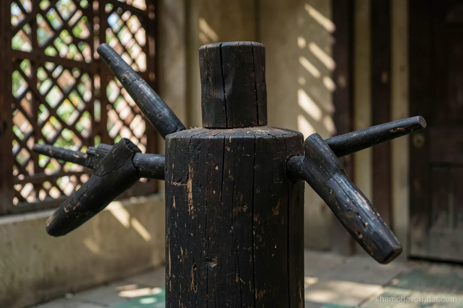 Close-up of a dark weathered wooden training dummy with three protruding arms in a quiet courtyard, morning light through a lattice window casting stripes, warm brown and jade palette